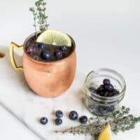 Blueberry lemonade Moscow Mule cocktail in a copper mule mug pictured among spare ingredients on a marble board with a neutral background.