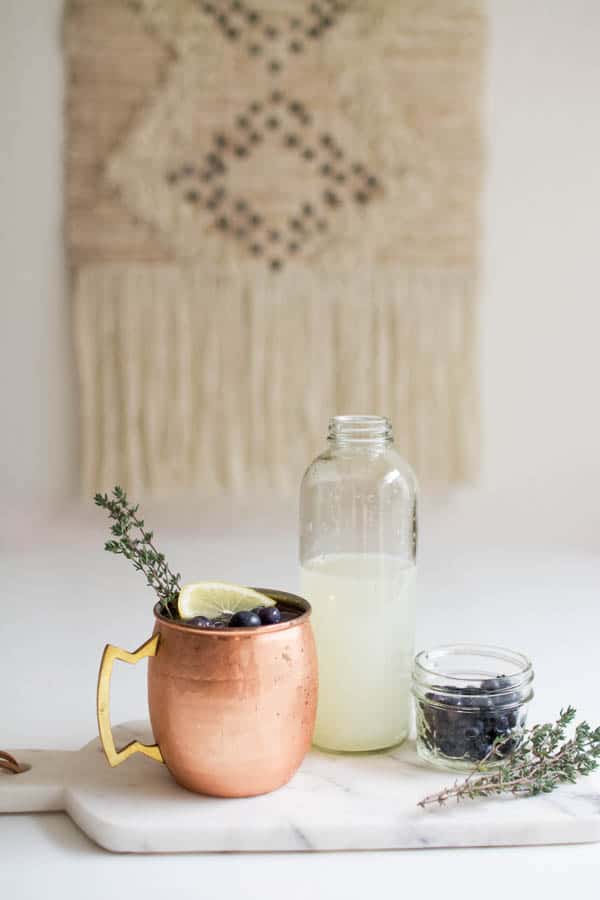 Copper Moscow Mule mug on a cutting board next to a jar of lemonade and a glass bowl of fresh blueberries and sprigs of fresh thyme.