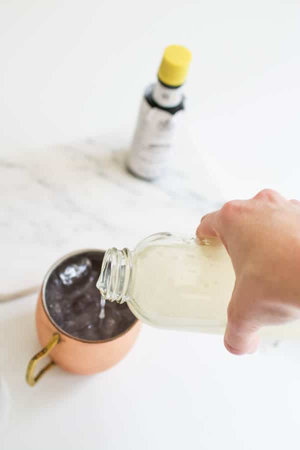 Hand pouring lemonade into a copper Moscow Mule mug with a bottle of cocktail bitters.