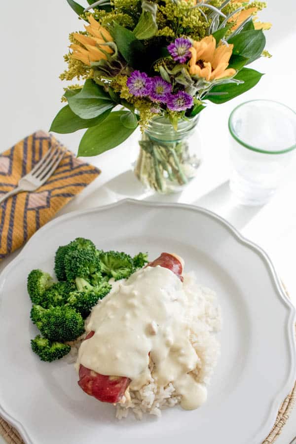 Baked chicken breast on top of rice on a dinner plate covered with a cream sauce next to a side of broccoli. 