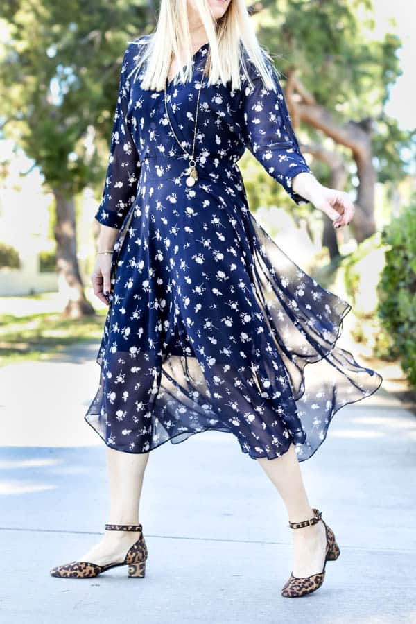 Blonde women in a blue floral dress outside with trees behind her. 