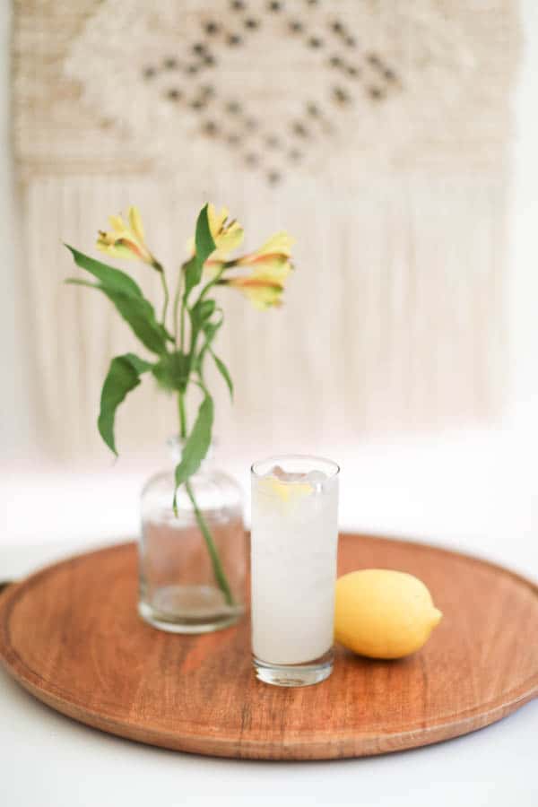 Tall cocktail glass on a wood tray on a table with a lemon next to it.