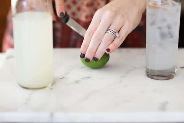 Woman cutting a lime on a cutting board with a cocktail glass next to her and a bottle of lemonade.