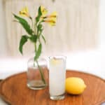 A tall cocktail glass on a wooden tray next to a yellow flower and lemon.