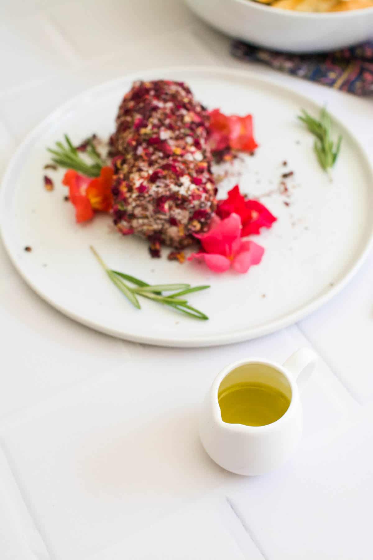 A spice crusted goat cheese log appetizer on a plate garnished with edible flowers with a small pitcher of olive oil next to it on a table.