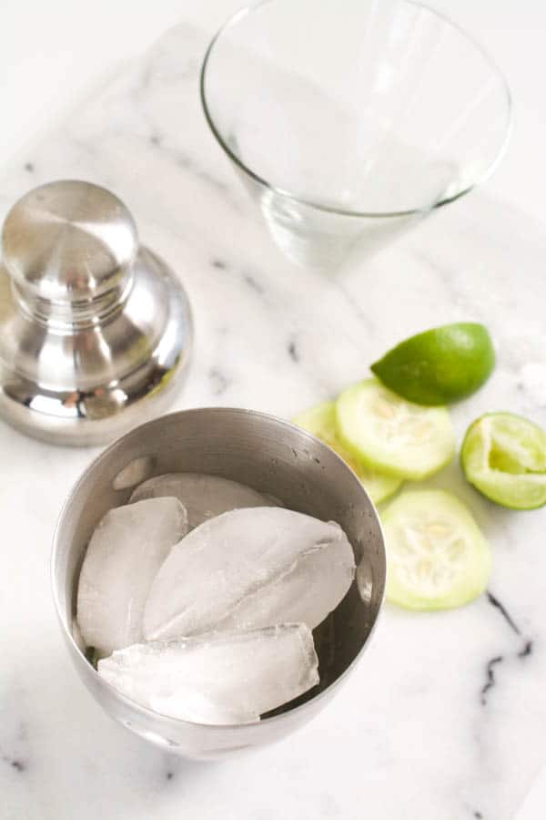 Cocktail shaker with ice in it next to limes and cucumbers slices.