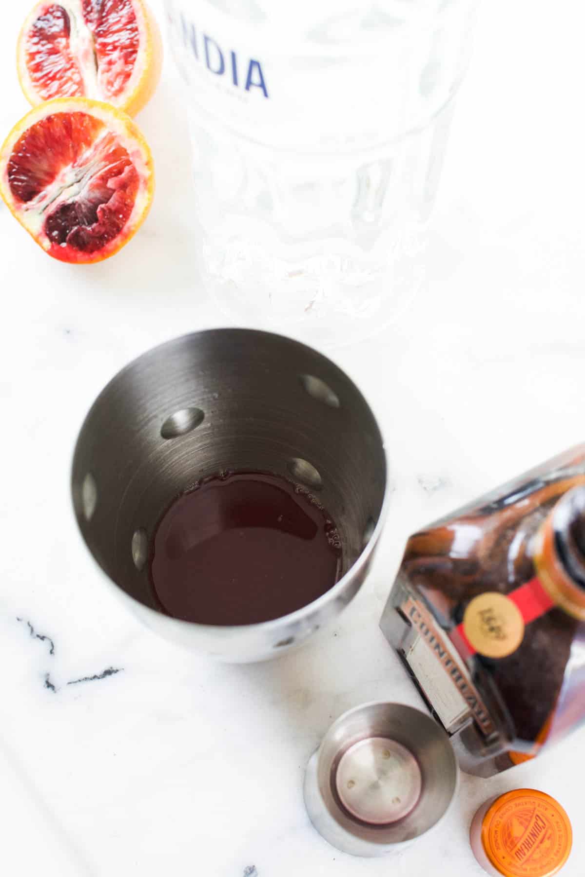 A cocktail shaker on a counter next to a bottle of Cointreau and cut blood oranges.