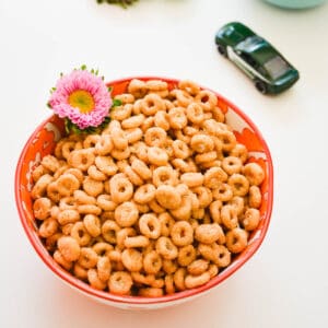 close up of an orange bowl with Cheerios, a small flower and a toy car in the background.