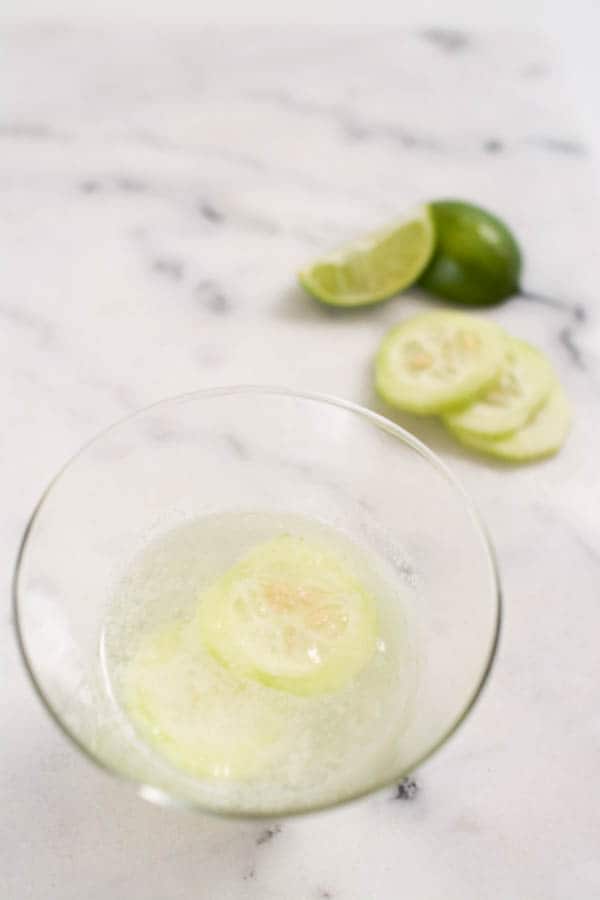 A cucumber slice garnish in the top of a cocktail in a glass on a table. 