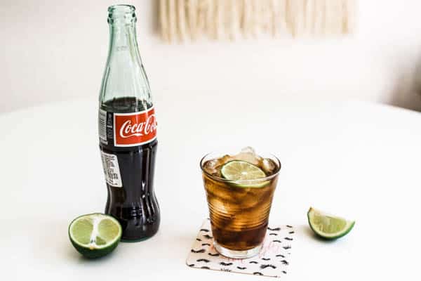 Glass of coke with ice and lime slice on a coaster on a table with a bottle of coke next to it and lime wedges.