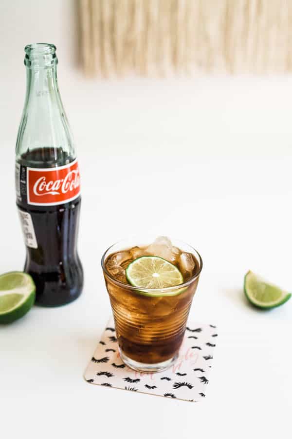 Glass of coke next to a coke bottle on a table with lime wedges around it.