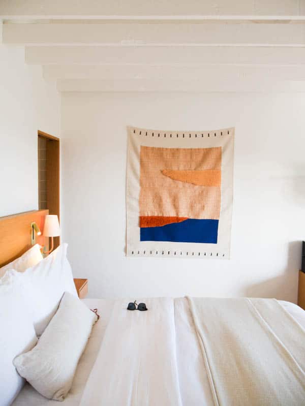 A guest room of Hotel Joaquin showing the bed with sunglasses laying on it and a textured wall hanging.
