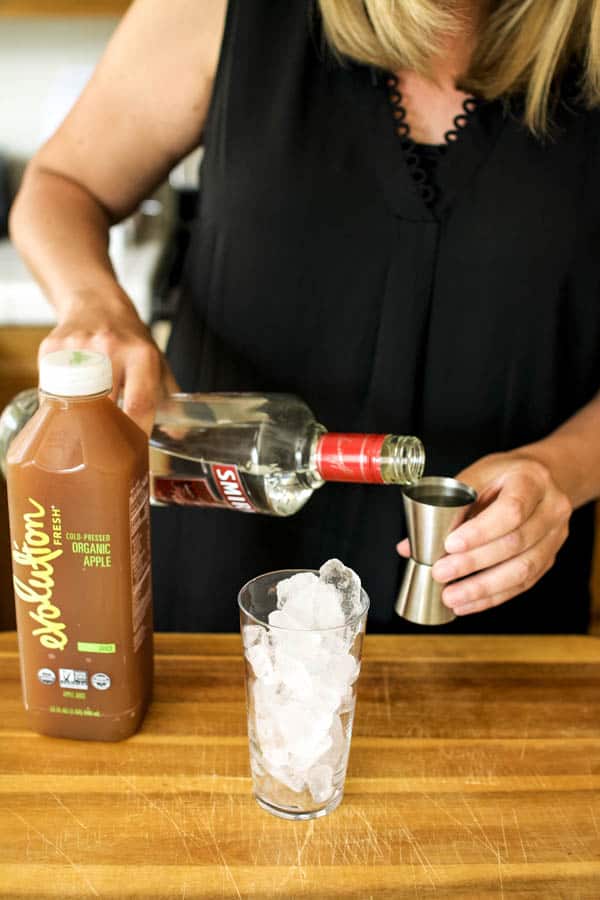 Girl adding vodka to a glass of ice with a bottle of apple juice sitting next to it.