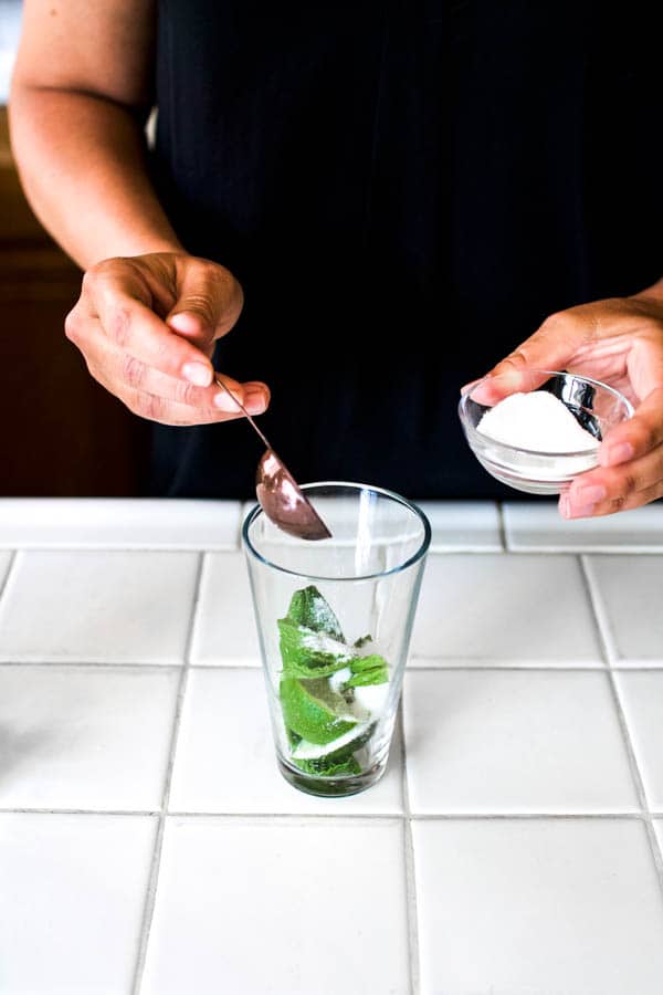Woman adding sugar to a glass with mint and limes for a mojito.