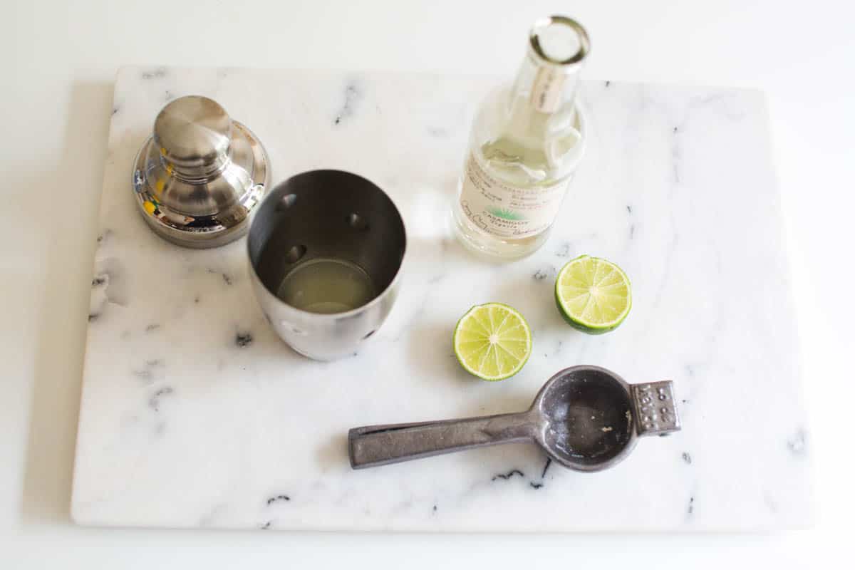 A cocktail shaker, citrus squeezer and cut limes on a marble cutting board next to a bottle of tequila.