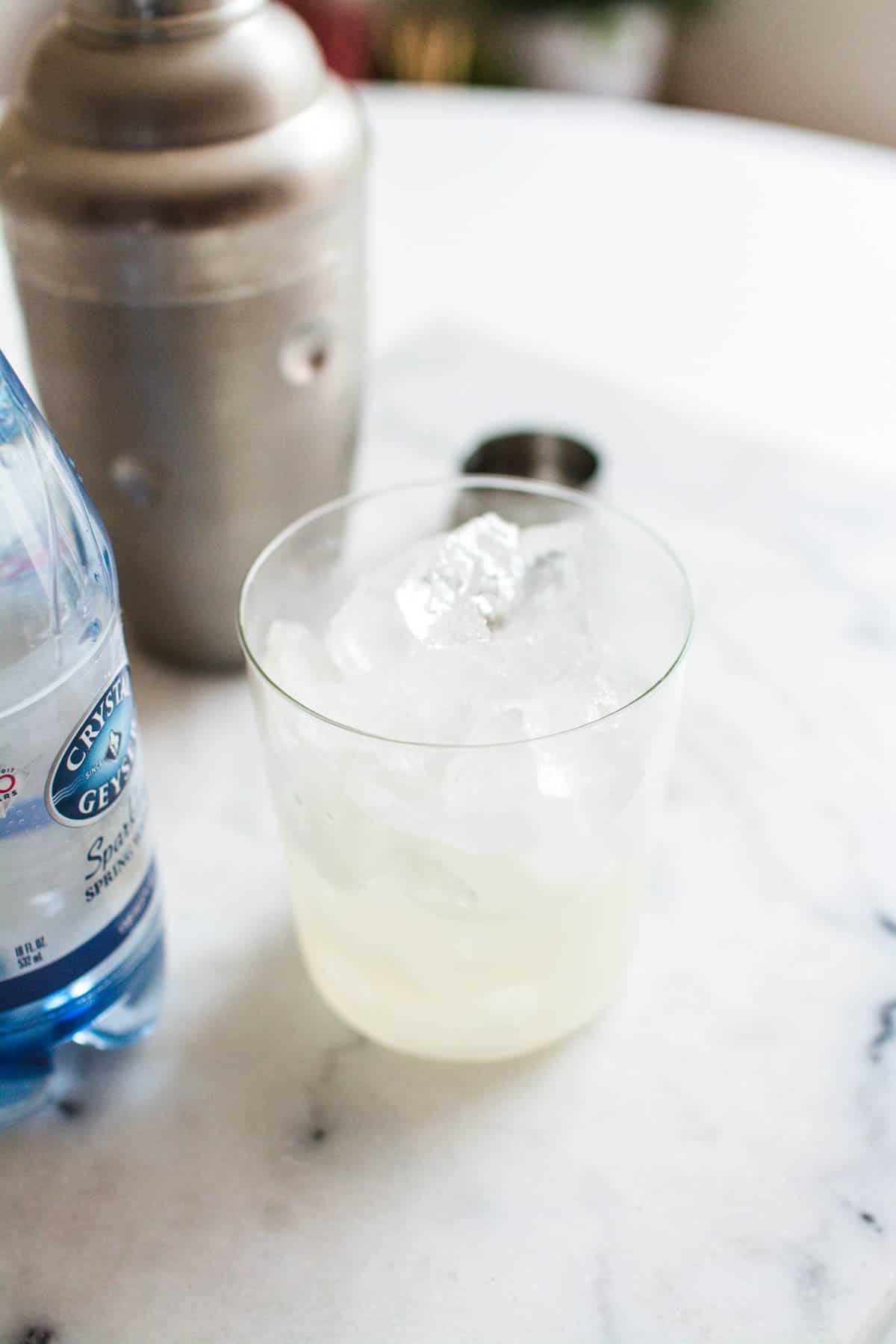 A cocktail glass on a marble cutting board with fresh lime juice and tequila next to a cocktail shaker.