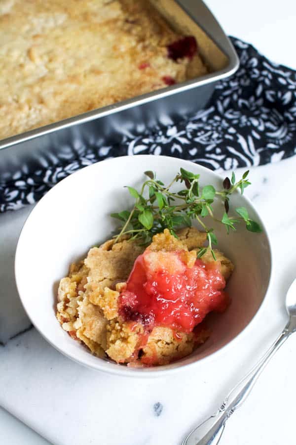 white bowl with strawberry pineapple dump cake and thyme sprigs on a table next to a pan of dump cake.