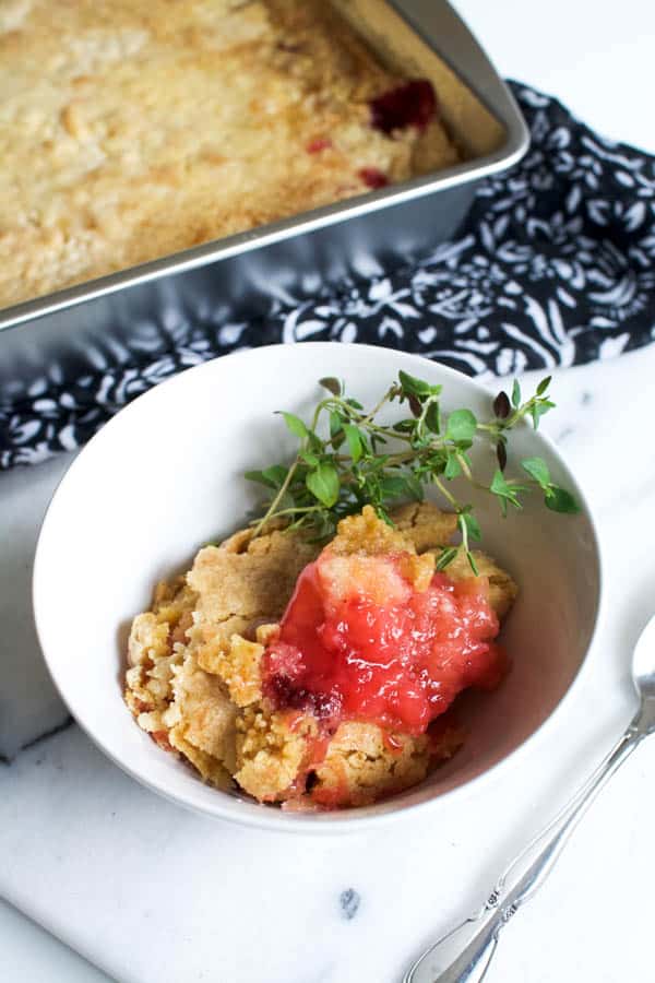 White bowl with strawberry pineapple dump cake and thyme sprigs on a table next to a pan of dump cake.