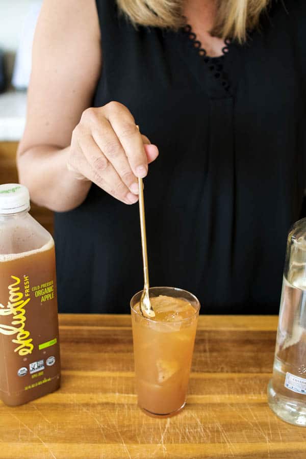 Woman stirring a vodka and apple juice 2 ingredient cocktail in a glass on a cutting board.