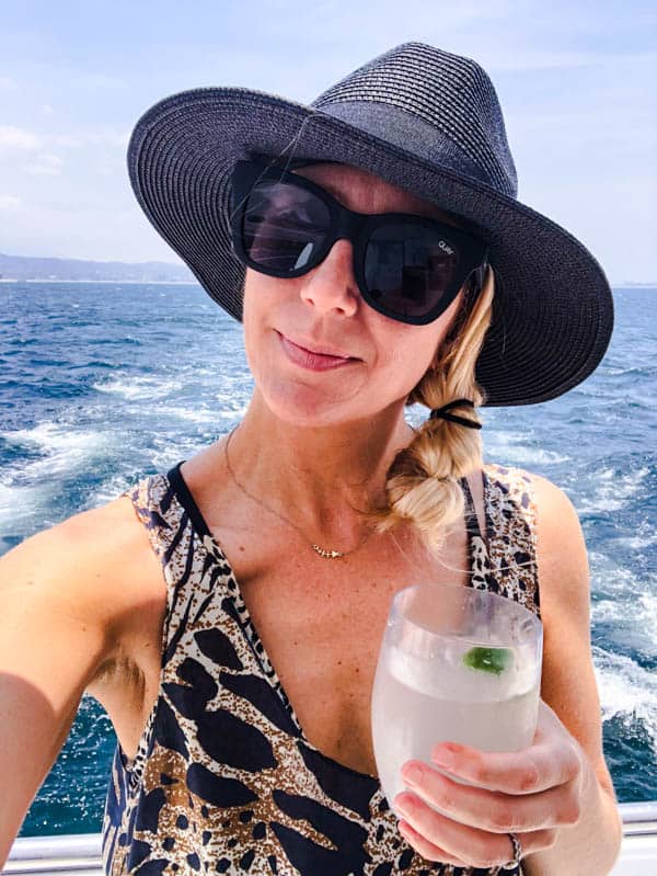 Women in black hat holding a cocktail with the ocean behind her.