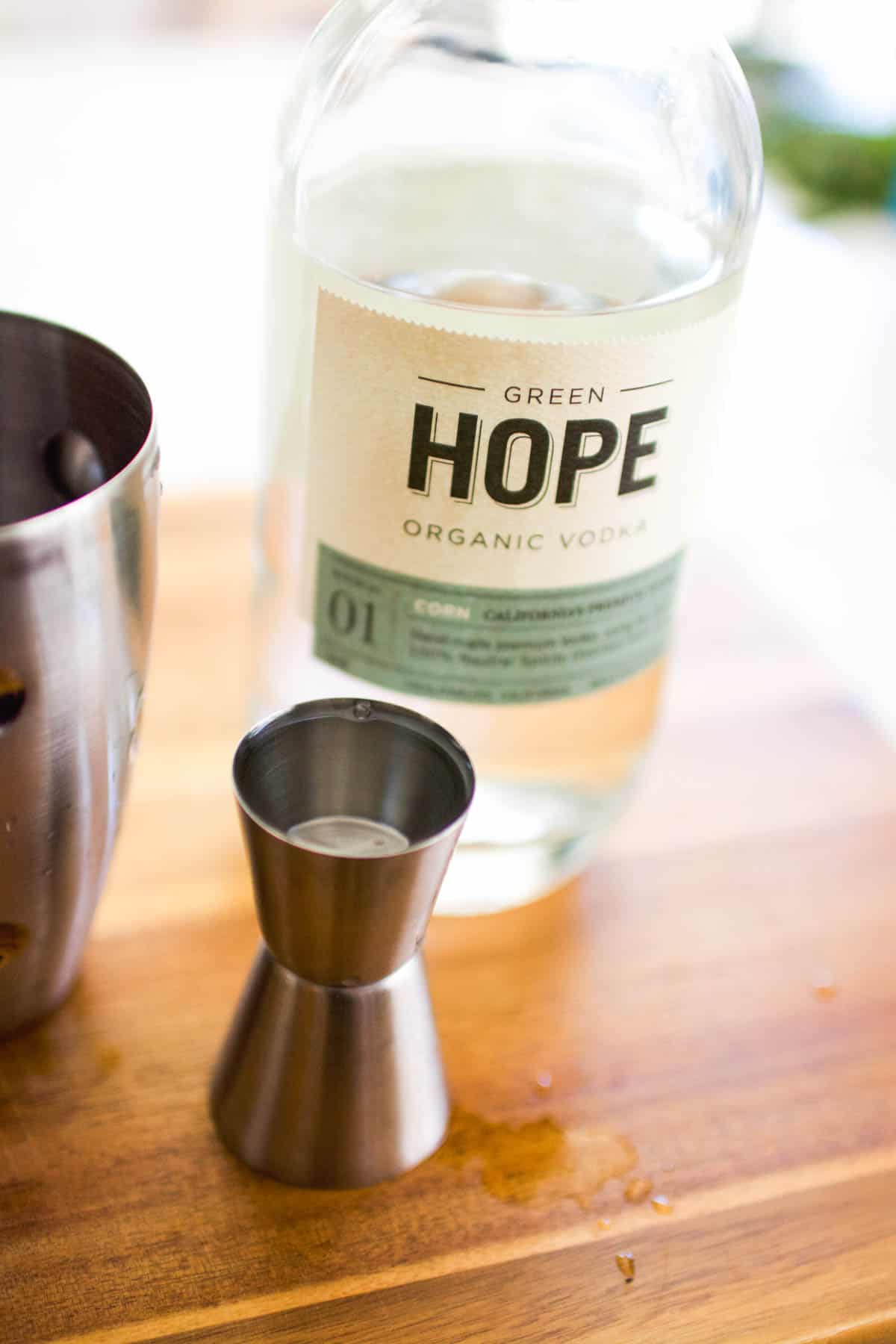 A jigger with vodka on a cutting board with a bottle of vodka behind it.