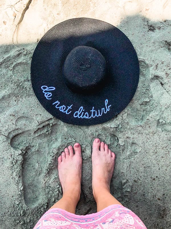an overhead shot of a hat that says do not disturb and feet in the sand on a beach