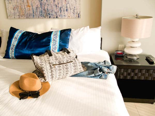 Carry on bag on a hotel bed next to a jean jackets, hat and sunglasses. 