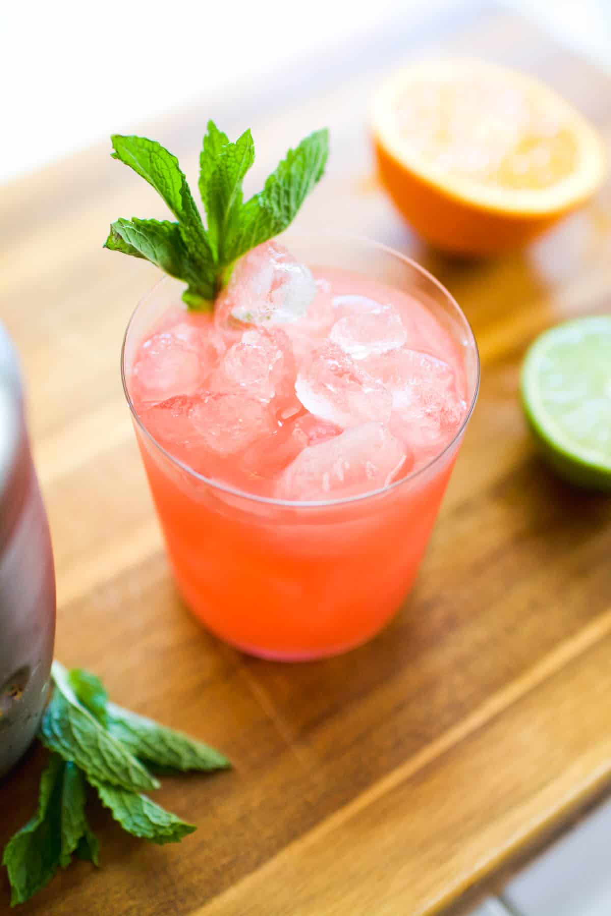 A pink cocktail with mint garnish on a cutting board next to cut citrus.