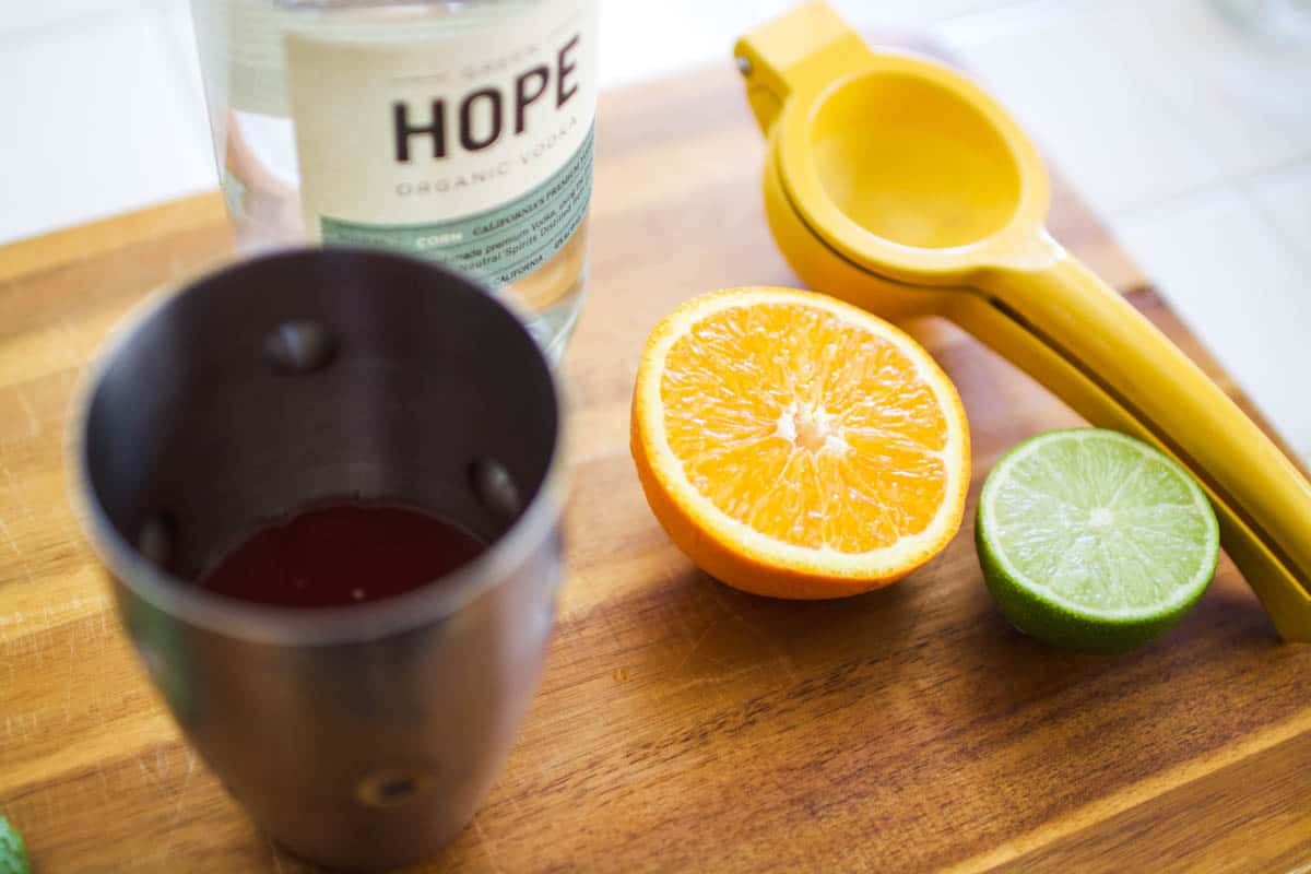 Half of an orange and lime on a cutting board next to a citrus squeezer and cocktail shaker.