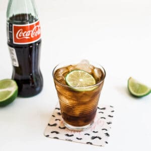 Close up of a cup holding coke with a lime garnish next to a bottle of Coke on the table.