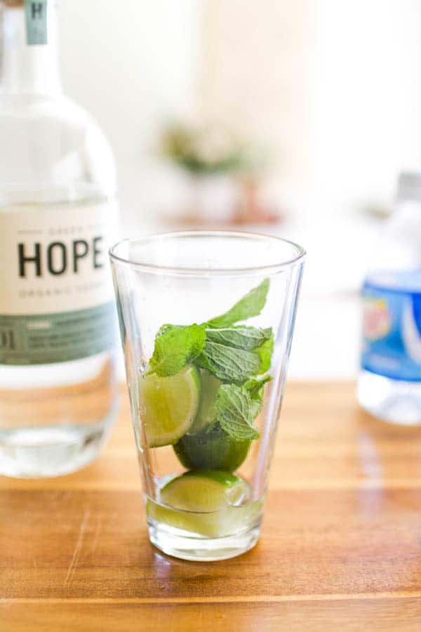 Lime wedges and mint in a glass next to a bottle of vodka and club soda