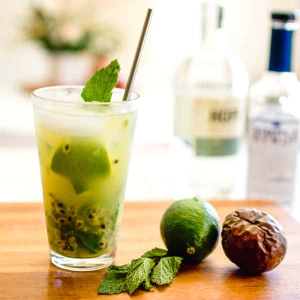 A finished passion fruit mojito on a cutting board next to a lime, mint and passion fruit