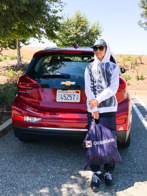 Teen boy leaning against the back of a Chevy Bolt holding a Crossroads Trading bag.