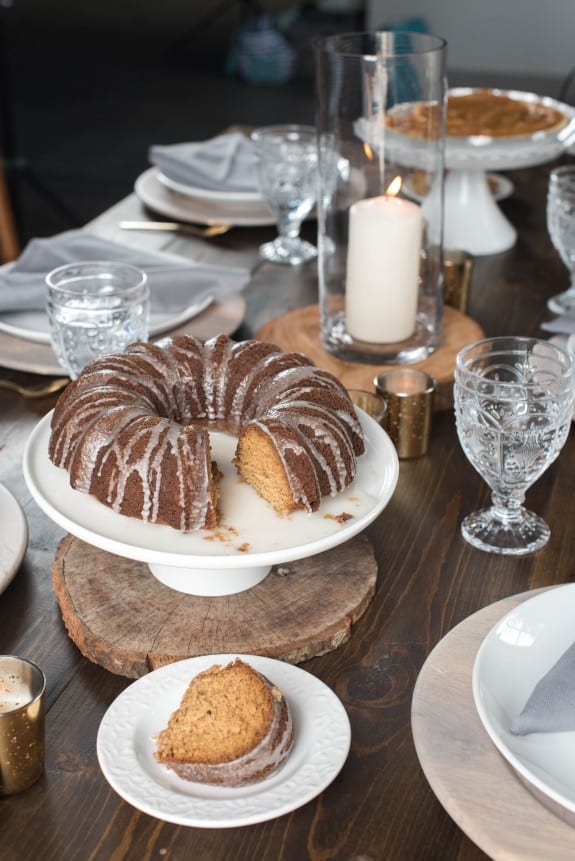 Slice of bundt cake with nutmeg on a small white dessert plate on a table next to a white cake plate on a party table. 