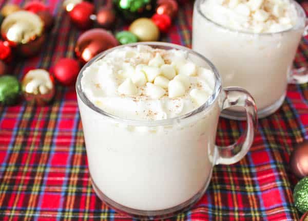 2 glass mugs on a red plaid covered table with white chocolate hot chocolate topped with whipped cream cinnamon and white chocolate chips. 