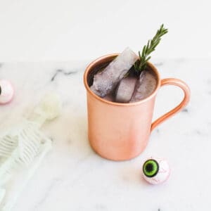 Close up of a copper mug with a Blackberry Moscow Mule in it with a sprig of fresh rosemary and an eyeball next to it.
