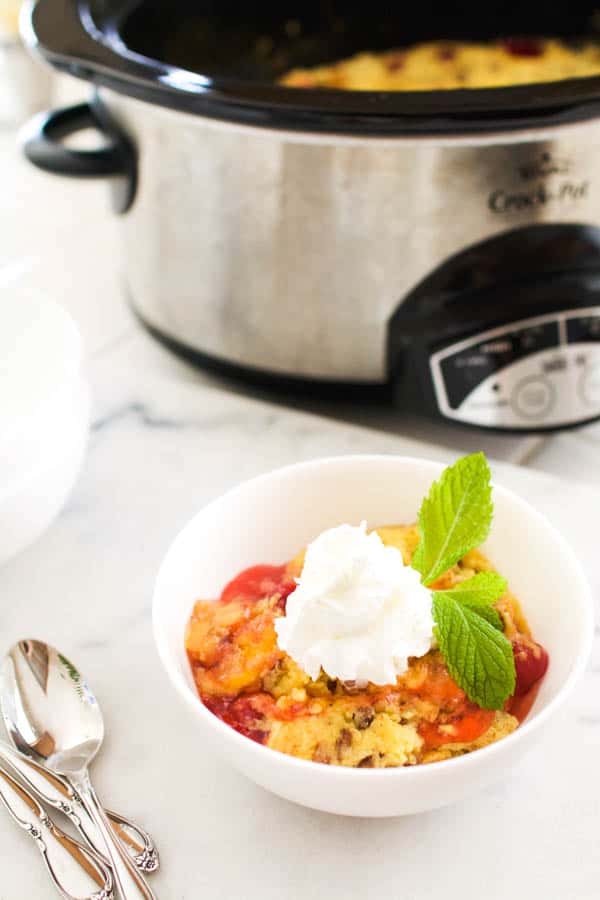 Apple Cherry dump cake with pecans and cherries in a bowl topped with whipped cream in front of a slow cooker. 