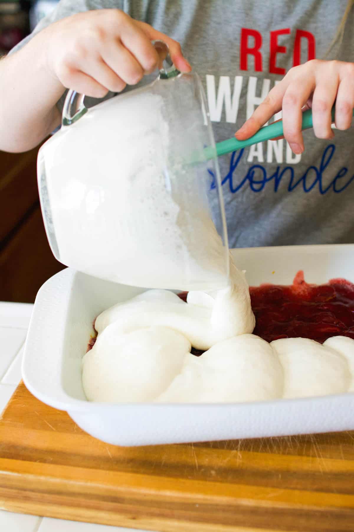 A woman pouring angel food cake batter on top of strawberry pie filling in a baking dish.