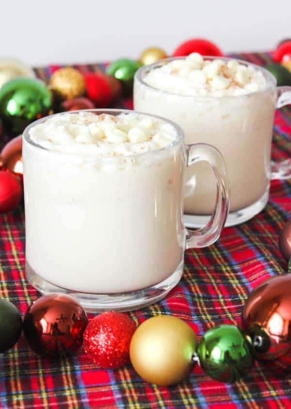 2 clear mugs holding white hot chocolate topped with whipped cream and white chocolate chips on a red plaid table cloth with Christmas ornaments in the background.