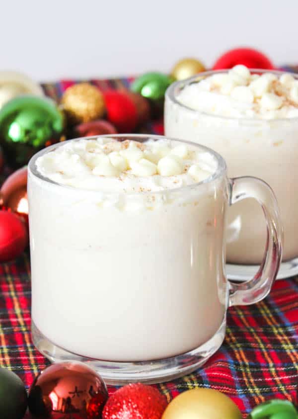 White hot chocolate recipe for cold weather served in 2 clear class mugs on a red plaid table with Christmas ornaments around the mugs. 