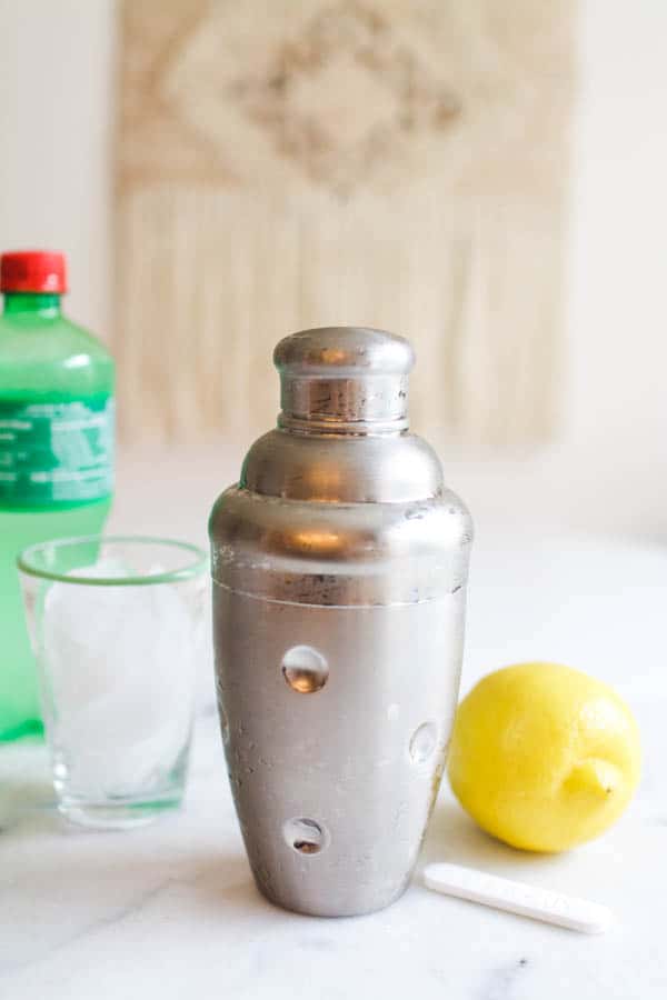 Cocktail shaker with a lemon, glass and bottle of Sprite next to it on a cutting board. 