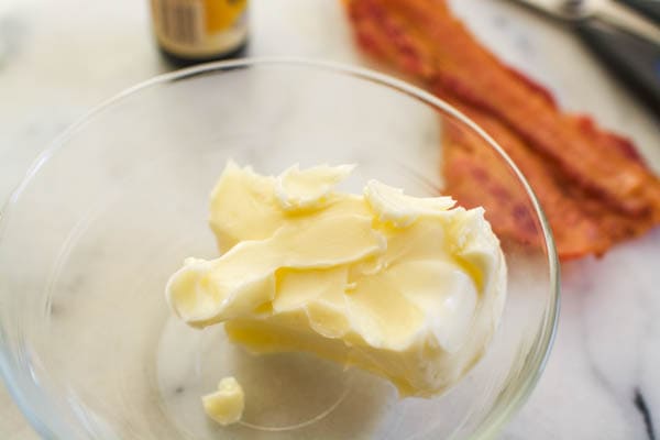 Softened butter for bacon butter in a glass bowl next to a few pieces of cooked bacon.