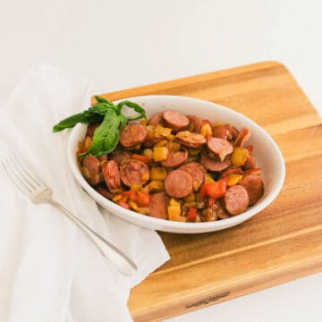 A bowl of sausage, onions and peppers on a cutting board with a basil garnish.