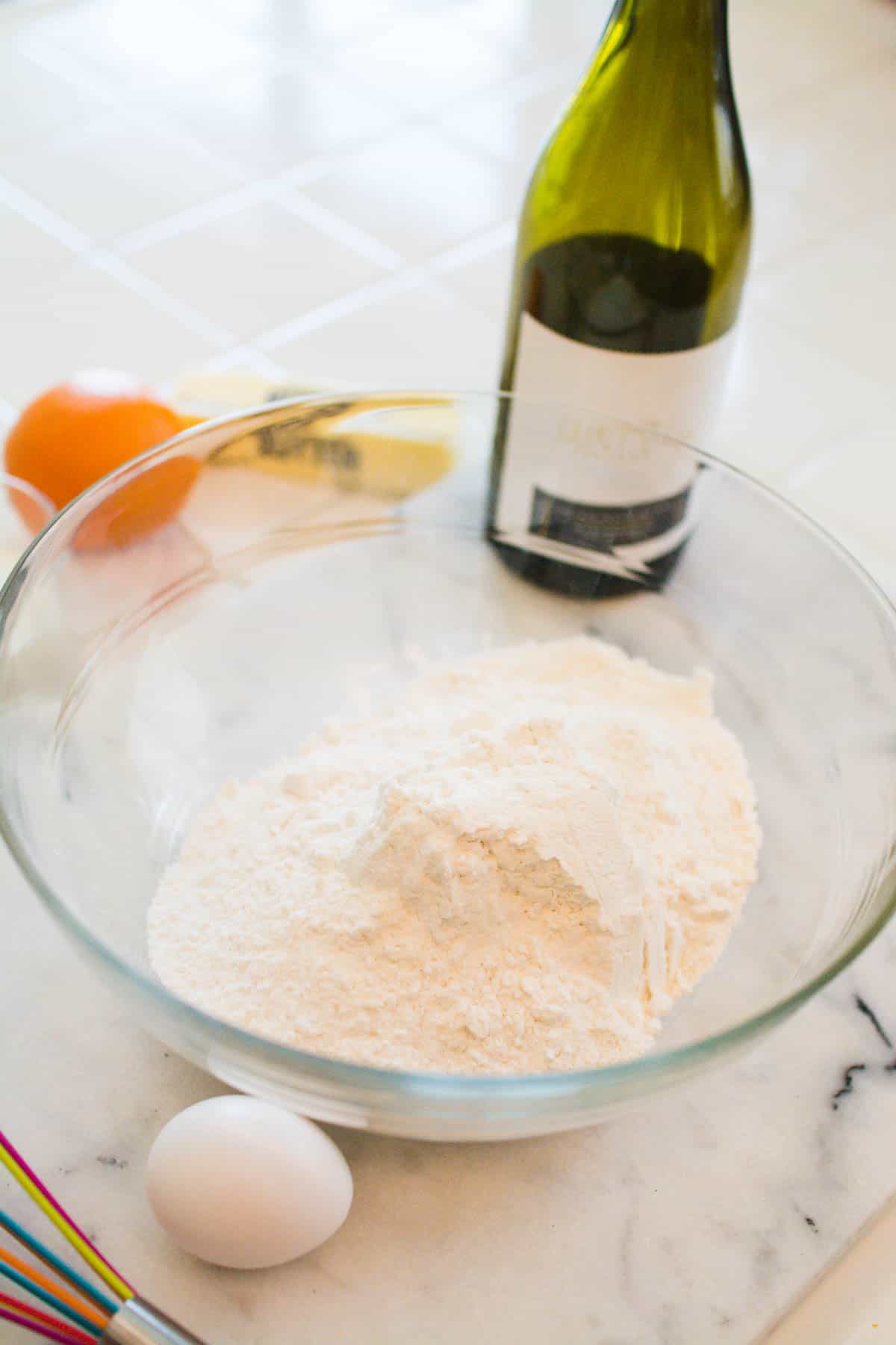 A glass bowl on a counter with boxed cake mix powder in it with an egg and bottle of wine next to it.