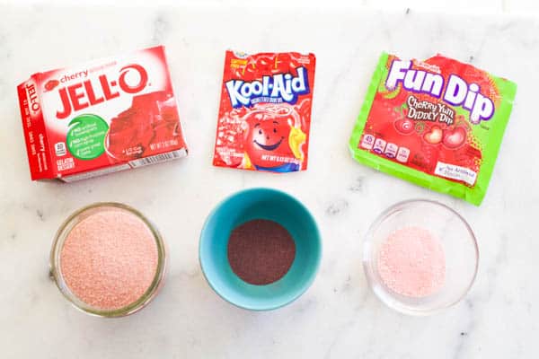 A box of jello next to a bowl with the Jello powder, a packet of KoolAid next to a bowl with the powder and a pack of Fun Dip candy next to a bowl of powder. 
