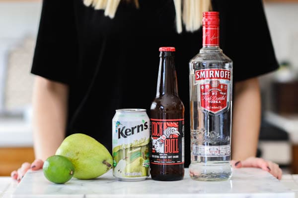 Ingredients for a spiced pear moscow mule on a counter with a woman standing behind it. 