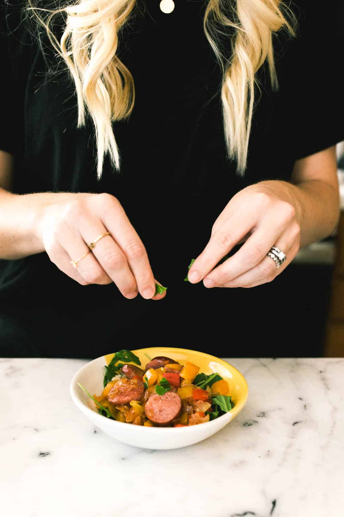 A woman adding fresh basil to a sausage bowl with quinoa to a bowl on the counter.