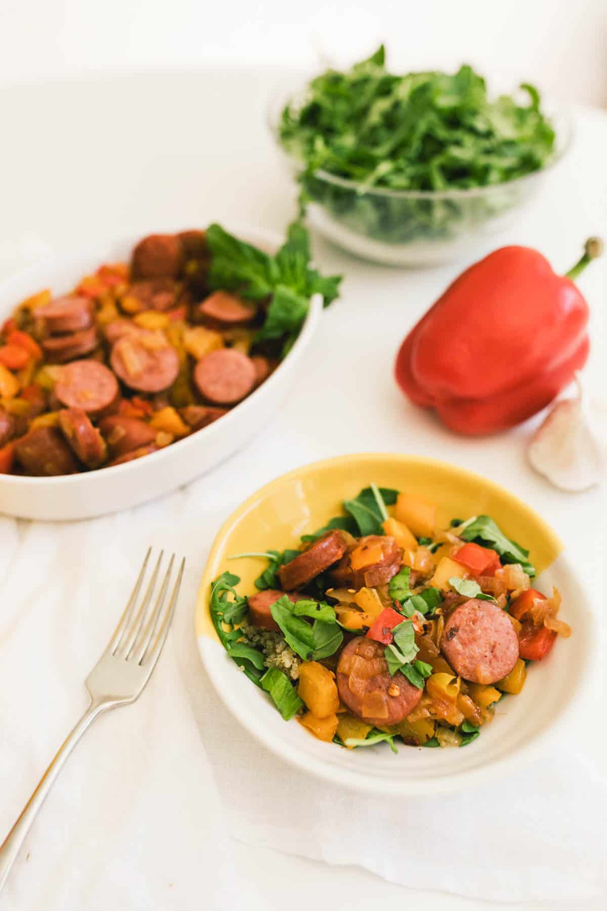 A serving dish and bowl of Quinoa and Sausage with peppers on a table next to a bowl of arugula and a red bell pepper.
