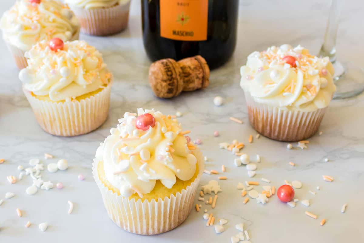 Frosted Prosecco cupcakes on a table that are topped with sprinkles next to a bottle of champagne. 