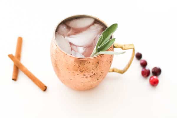 A spiced Cranberry Mule for the holidays in a copper mug with a sage garnish and cinnamon and cranberries on the table next to it.